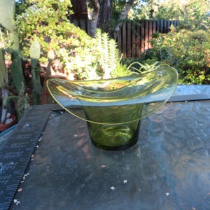 May include: A translucent olive green glass bowl featuring a wide, flared, and undulating rim that creates a distinct, organic shape. The glass is smooth and clear, allowing light to pass through the vessel, which sits on a flat base.