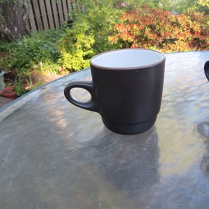 May include: A dark brown coffee mug with a white interior and a rounded handle. The mug has a simple, classic design and is made of ceramic. The mug is sitting on a glass table outside.