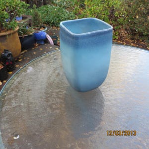 May include: A blue ceramic vase with a square top and rounded base. The vase has a gradient color scheme, transitioning from a dark blue at the top to a lighter blue towards the bottom. The vase is approximately 10 inches tall.