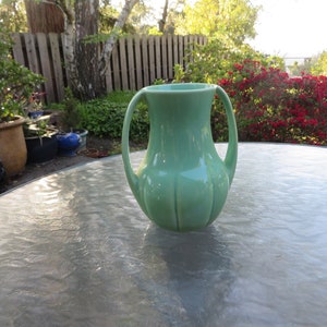 May include: A light green ceramic pitcher with two curved handles. The pitcher has a ribbed design on the body and sits on a glass table. The background includes a garden with plants and a wooden fence.