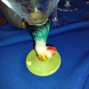 May include: A clear martini glass with a green and white rooster-shaped base. The rooster is perched on a round, light green base. The glass has a triangular bowl and is set against a blue background.
