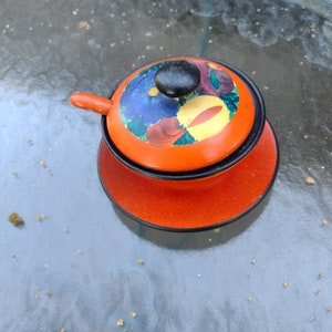 May include: An orange enamelware serving dish with a lid and a small handle. The lid features a floral design with blue, red, and yellow accents. The dish sits on a matching orange saucer.