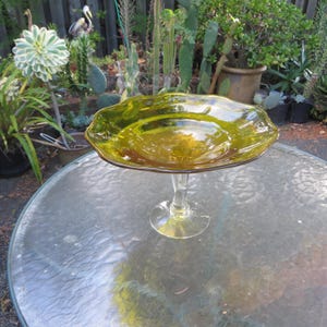 May include: A pedestal bowl made of translucent olive green glass with a wide, scalloped rim. The bowl is supported by a clear, twisted glass stem that connects to a circular, flat base, providing a stable foundation for the elevated dish.