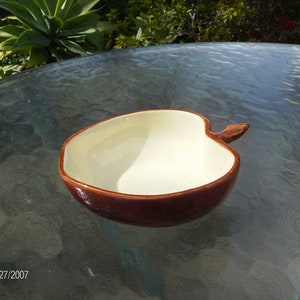 May include: Ceramic apple-shaped bowl with a brown exterior and a cream-colored interior. The bowl has a stem-like handle and is likely used for serving or display. The bowl is sitting on a glass table.