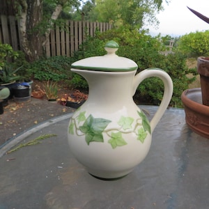 May include: Cream-colored ceramic pitcher with a lid and handle. The pitcher is decorated with green ivy leaf designs. The lid and base have green trim. The pitcher is sitting on a glass table outdoors.