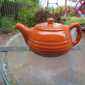 May include: A ceramic teapot with a lid and handle. The teapot is a warm orange color with horizontal ridged detailing around the body. The spout and handle are curved. The teapot is sitting on a glass table.