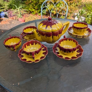 May include: A vintage yellow and burgundy ceramic tea set. The set includes a teapot, cups, and saucers. The teapot has a lid with a red knob. The cups and saucers have a scalloped edge design. The tea set is on a glass table.