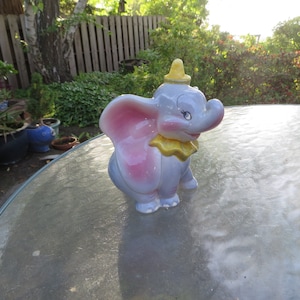 May include: A ceramic figurine of Dumbo the elephant. The figurine is primarily gray with pink ears, a yellow collar, and a yellow hat. The elephant is in a seated position with a smiling expression.