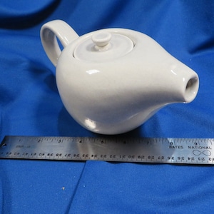 May include: A white ceramic teapot with a rounded body, a curved handle, and a spout. The lid has a small knob. A ruler is placed next to the teapot. The teapot is approximately 6 inches long.