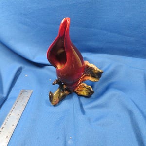 May include: A ceramic sculpture of a maroon-colored flower with a dark interior and a textured, multi-colored base. The flower has a curved, open shape. The base has green, brown, and yellow tones. The sculpture is on a blue surface.