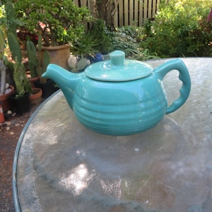 May include: A turquoise ceramic teapot with a matching lid and handle. The teapot has horizontal ridges around its body and sits on a glass table. The background features greenery and potted plants.