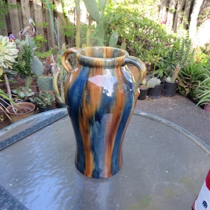 May include: A ceramic vase with a glossy finish featuring vertical streaks of deep blue, amber, and brown glaze. The vessel has a wide, rounded body, a flared rim, and two symmetrical handles attached to the upper sides of the neck.