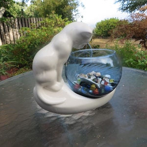 May include: A white ceramic cat figurine with a curved base, supporting a clear glass bowl. The bowl contains colorful stones. The cat is posed looking down into the bowl. The piece is likely a decorative item.