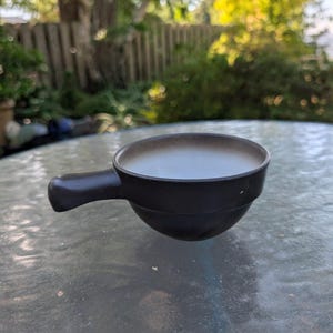 May include: A small, dark brown ceramic bowl with a handle. The bowl has a rounded shape and a light brown interior. The handle is attached to the side of the bowl. The bowl is sitting on a glass table.