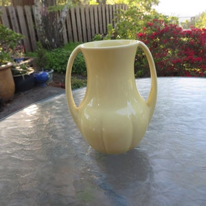 May include: A pale yellow ceramic vase with two curved handles. The vase has a rounded base and a flared top. The vase is sitting on a glass table outdoors, with a garden in the background.