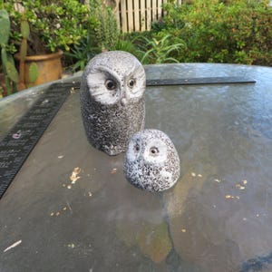 May include: Two owl figurines with a speckled gray and white finish. The larger owl stands upright with prominent facial disks, while the smaller owl is rounded and compact. Both feature dark, circular eyes and are positioned near a black ruler.