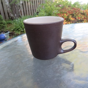 May include: A brown ceramic mug with a beige interior. The mug has a simple, tapered design and a curved handle. The mug is sitting on a glass table outdoors.
