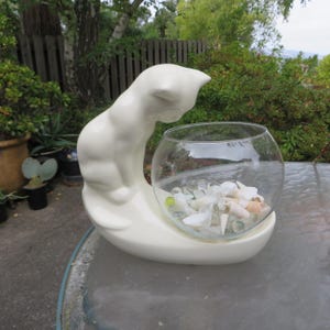 May include: A white ceramic cat-shaped planter with a clear glass bowl. The cat is perched above the bowl, which contains seashells and small glass beads. The planter sits on a curved base.