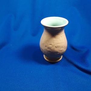 May include: A small, beige vase with a textured surface and a light green interior. The vase has a rounded body and a flared rim. The base is a lighter shade of beige. The vase is set against a blue background.
