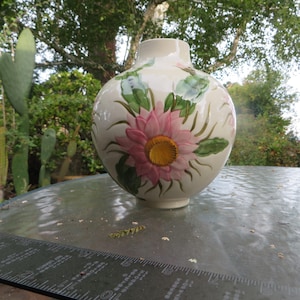 May include: A cream-colored ceramic vase with a rounded body and a narrow neck, featuring a hand-painted pink flower with a yellow center and green leaves. A black ruler is positioned in the foreground, showing a measurement of 6 inches.