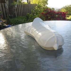 May include: A white, abstract sculpture resembling a stylized shoe or foot, resting on a reflective surface. The sculpture has a smooth, glossy finish and is positioned outdoors with a blurred background of greenery and a wooden fence.