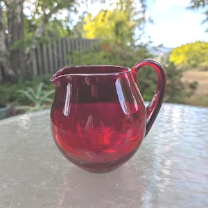 May include: A small, round, ruby-red glass creamer with a handle and a pouring spout. The creamer is sitting on a glass table outside, with a blurred background of trees and a fence.