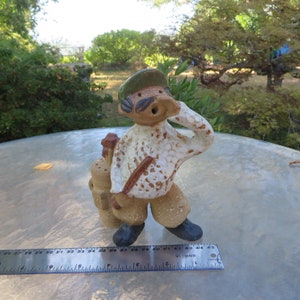 May include: A ceramic figurine of a golfer in a white shirt, khaki pants, and a green cap. The golfer is holding his hand up to his forehead and carrying a golf bag. The figurine is approximately 4 inches tall.
