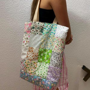 Puede incluir: Un bolso tote rectangular construido a partir de un patchwork de varios cuadrados de tela con estampados florales y diversos diseños. El bolso presenta una mezcla de colores verde, rosa, azul y blanco con diversos estampados botánicos y dos asas de tela beige.