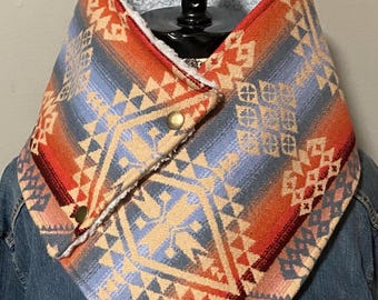 Puede incluir: Una bufanda tipo cuello con un patrón geométrico en tonos naranja, azul y crema. La bufanda está forrada con forro polar sherpa blanco y asegurada con dos broches de metal dorado. Se lleva sobre una chaqueta de mezclilla azul.