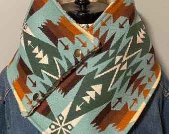 Puede incluir: Un calentador de cuello tipo cowl con estampado turquesa, marrón y blanco con forro polar. El calentador de cuello tiene un diseño geométrico con formas de flechas y triángulos. Se asegura con dos botones color bronce.