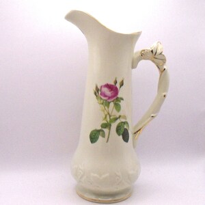 May include: White porcelain pitcher with a gold trim and a floral design featuring a pink rose with green leaves. The handle is shaped like a branch with a gold trim.