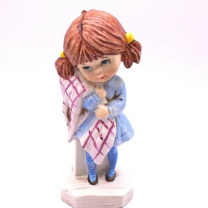May include: A porcelain figurine of a girl with brown hair and blue eyes. She is wearing a blue dress with a pink and white plaid pattern. She is holding a blanket in her arms and is standing on a white base.