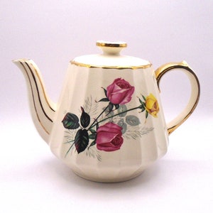 May include: A white teapot with a gold trim and a floral design featuring pink and yellow roses. The teapot has a fluted body and a gold lid.