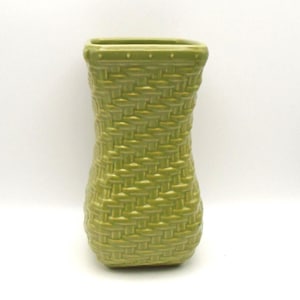 May include: A green ceramic vase with a woven basket design. The vase has a rectangular top and a slightly flared body. The color is a muted, earthy green, and the texture mimics the look of a woven basket. The vase is suitable for displaying flowers or as a decorative item.