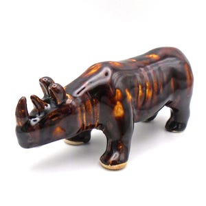 May include: A ceramic figurine of a rhinoceros in a glossy, dark brown glaze with lighter brown accents. The rhino is depicted in a walking pose, showcasing its horns and detailed features. The figurine is a decorative item.
