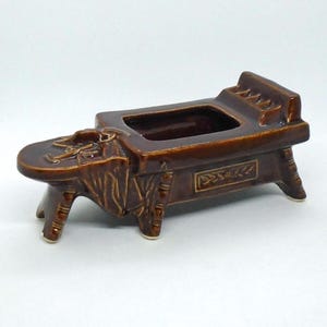 May include: A brown ceramic planter shaped like a small bed with four legs. The planter has a rectangular opening for planting and a decorative design on the side.