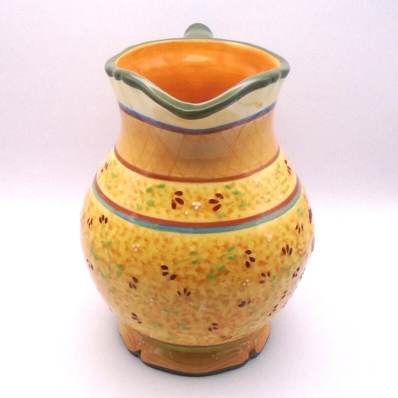 May include: A yellow ceramic pitcher with a green rim and floral pattern. The pitcher has a wide mouth and a handle.