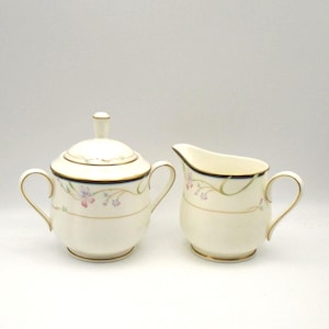 May include: Cream-colored sugar bowl with lid and creamer set. Both pieces have gold trim, dark blue bands, and delicate floral designs in pink and green. The sugar bowl has two handles.