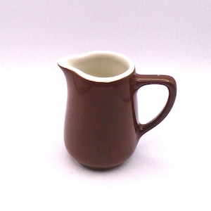 May include: A small brown ceramic creamer with a white rim. The creamer has a rounded body and a handle.
