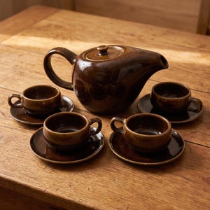 May include: A dark brown glazed ceramic tea set consisting of one teapot with a lid and four matching teacups, each resting on a small circular saucer. The set features a smooth, reflective finish and a uniform deep brown colour throughout.