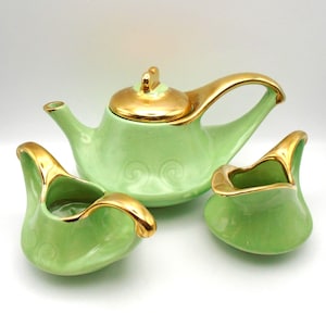 May include: A vintage tea set in a soft green color with gold trim. The set includes a teapot with a lid, and two small creamers. The teapot and creamers have a swirl design on the side.