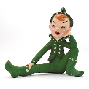 May include: A vintage figurine of a seated elf in a green outfit with white polka dots. The elf has red hair, blue eyes, and is resting their head on their hand. The figurine is on a white background.
