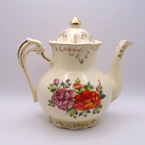 May include: A cream-coloured teapot with a floral design. The teapot has a gold trim and a gold lid with a floral design. The teapot is decorated with pink, orange, and yellow roses.