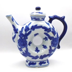 May include: A white ceramic teapot with a blue and white floral design. The teapot has a round body with a hole in the center and a lid. The teapot has a spout and a handle.