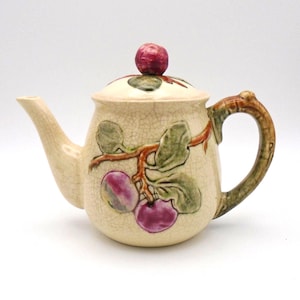 May include: A cream-coloured teapot with a crackled glaze and a floral design featuring purple plums and green leaves. The teapot has a brown handle and a lid with a single purple plum on top.