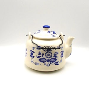 May include: Cream-colored ceramic teapot with blue floral patterns and a matching lid. The teapot has a black wire handle and a blue knob on the lid. The spout is curved, and the design is reminiscent of vintage tableware.