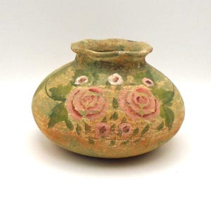 May include: A small, round, hand-painted ceramic vase. The vase is mainly green with pink roses and green leaves painted on the front. The rim and base have a weathered, aged appearance. The vase is likely for decorative purposes.
