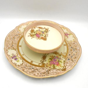 May include: A vintage teacup and saucer set. The cup and saucer are pale pink and cream with gold trim and floral designs. The saucer has a scalloped edge and intricate detailing. The cup is filled with a white liquid.
