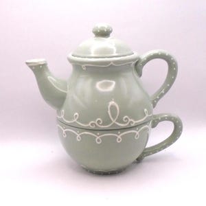 May include: A sage green teapot with a white lid and white decorative trim. The teapot has a matching cup with a handle.