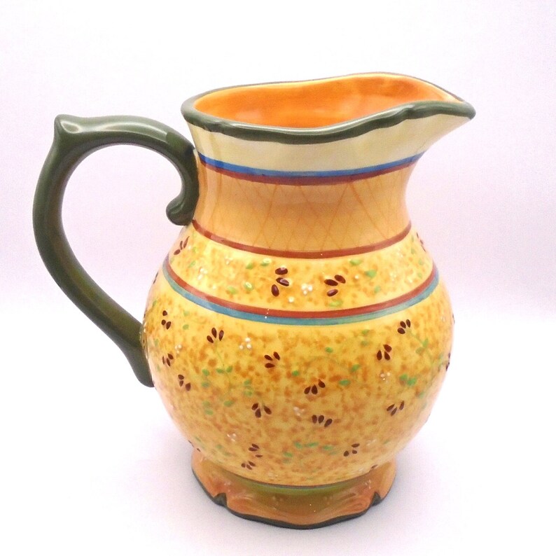 May include: A yellow ceramic pitcher with a green handle and a floral pattern. The pitcher has a scalloped rim and a wide base.